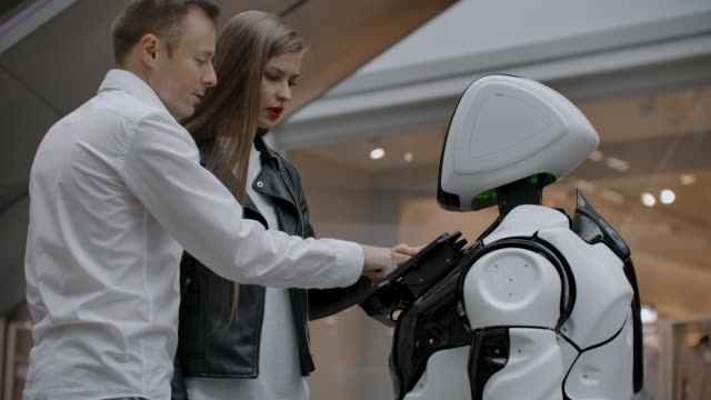 Ein-Mann-und-eine-Frau-in-der-Mall-interagieren-mit-einem-Roboterberater,-indem-sie-auf-den-Bildschirm-tippen-und-lächeln.-Cyborg-Android-hilft-Menschen.