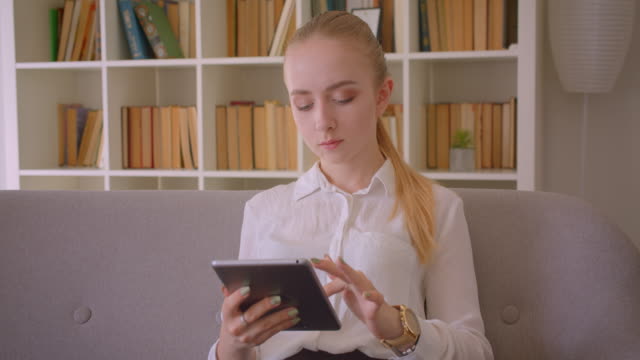 Nahaufnahme-Porträt-der-jungen-hübschen-kaukasischen-blonden-Studentin-mit-dem-Tablet,-das-auf-die-Kamera-schaut-und-fröhlich-auf-der-Couch-in-der-Wohnung-sitzt