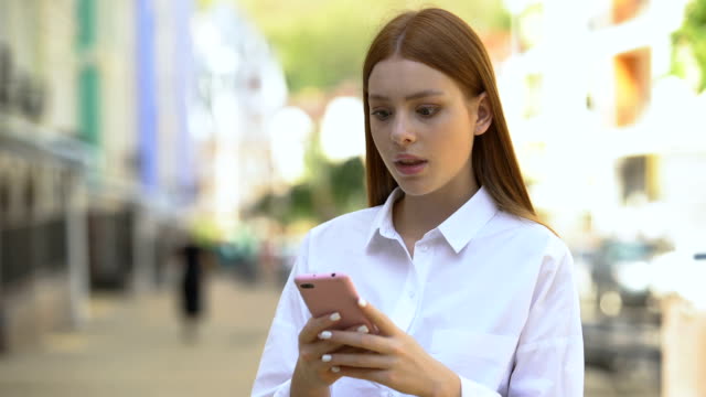 Young-redhead-female-scrolling-social-network-on-smartphone,-shocked-by-news