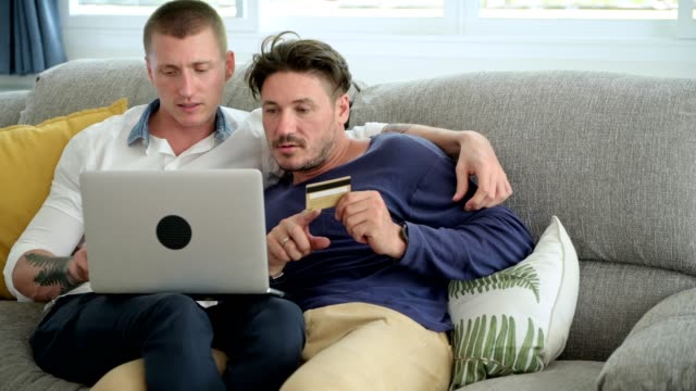 Gay-couple-relaxing-on-couch-using-laptop-computer.-Telling-what-to-key-into-laptop.