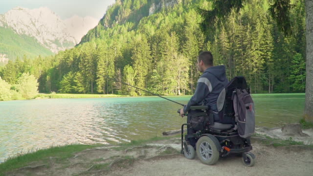 Cámara-lenta-de-pescador-discapacitado-en-una-silla-de-ruedas-eléctrica-pescando-en-un-hermoso-lago-cerca-del-bosque-y-la-montaña-en-la-parte-posterior