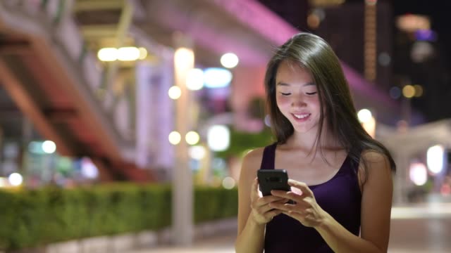 Glückliche-junge-schöne-asiatische-Frau-mit-Telefon-im-Freien-in-der-Nacht