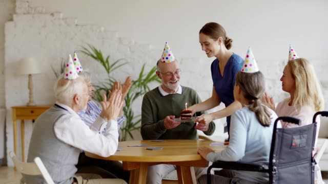 Tracking-shot-of-nurse-bringing-birthday-cake-for-elderly-man-playing-cards-with-aged-friends-sitting-at-table-in-nursing-home.-Happy-senior-man-getting-wishes,-kisses-and-hugs-and-blowing-candle-out