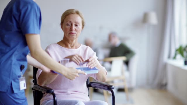Behinderte-ältere-Frau-sitzt-im-Rollstuhl-und-schaut-nachdenklich-weg.-Traurige-Patientin-schaut-in-die-Kamera,-wenn-junge-Krankenschwester-bringt-ihre-Vitaminpillen-und-Glas-Wasser