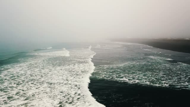 Aerial-view-of-ocean-shore-and-waves-with-surfers-in-fog