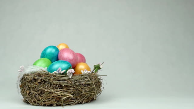 Chicken-colorful-eggs-in-a-nest-on-a-white-background-in-which-falls-a-light-feather,-Easter,-copy-space,-slow-motion,-traditional