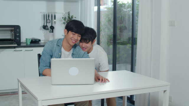 Young-Gay-couple-using-computer-laptop-at-modern-home.-Asian-LGBTQ-men-happy-relax-fun-using-technology-watching-movie-in-internet-together-while-sitting-table-in-kitchen-at-house-concept.