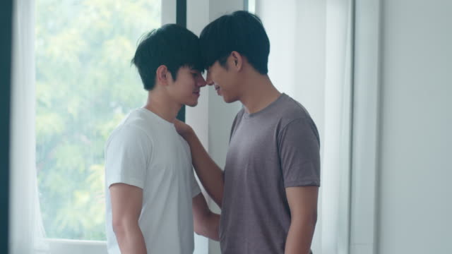Asian-Gay-couple-standing-and-hugging-near-the-window-at-home.-Young-Asian-LGBTQ+-men-kissing-happy-relax-rest-together-spend-romantic-time-in-living-room-at-modern-house-in-the-morning-concept.