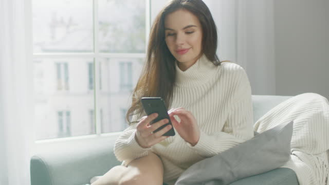 Beautiful-Young-Woman-Using-Smartphone-Smilingly,-while-Sitting-on-the-Chair.-Sensual-Girl-Wearing-Sweater,-Surfs-Internet,-Posts-on-Social-Media-while-Relaxing-in-Cozy-Apartment.-Closeup-Portrait