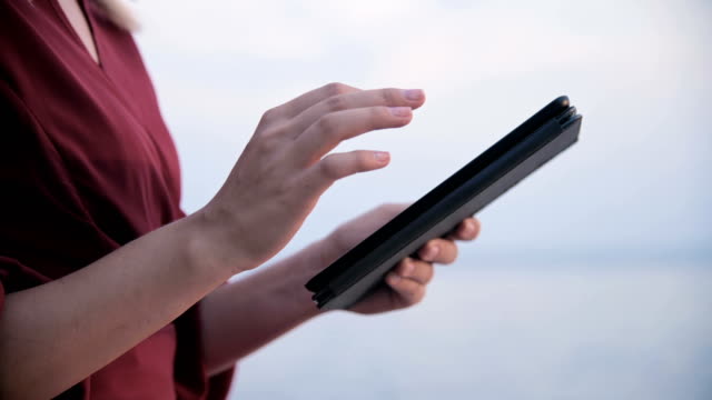 Close-up-An-attractive-young-girl-hands-in-a-summer-red-dress-sits-on-a-stone-by-the-sea-in-the-evening-and-looks-at-something-on-a-tablet.-Swipe-across-the-screen