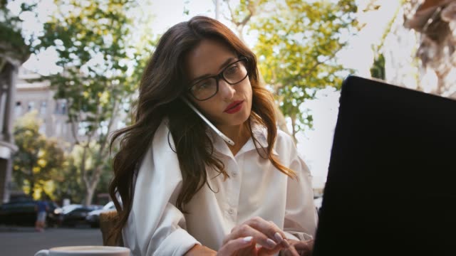 Business-woman-in-glasses,-white-shirt.-Sitting-at-table-with-cup-of-coffee-and-laptop-in-outdoor-cafe.-Talking-on-cellphone,-typing.-Slow-motion