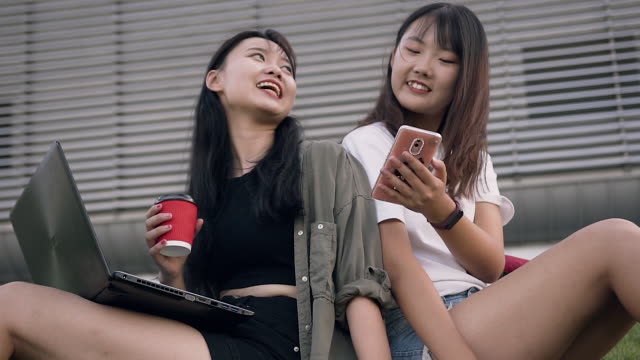 Hermosas-chicas-asiáticas-sonrientes-felices-sentadas-cerca-de-un-gran-edificio-urbano-y-usando-el-teléfono-para-ver-la-foto