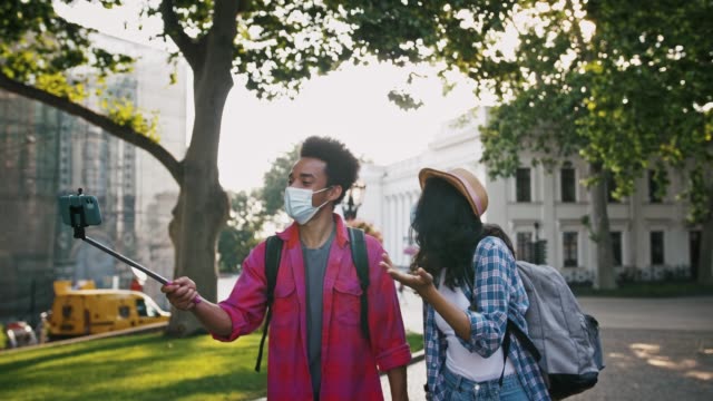 Afro-american-pair-in-protective-masks-recording-video-for-subscribers-on-cellphone-while-walking-along-city-street