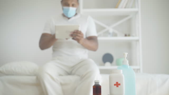 Close-up-of-sanitizer-and-pills-with-blurred-African-American-man-using-tablet-at-the-background.-Ill-male-patient-in-face-mask-sitting-on-bed-in-hospital-ward-surfing-Internet.-Covid-19-pandemic.