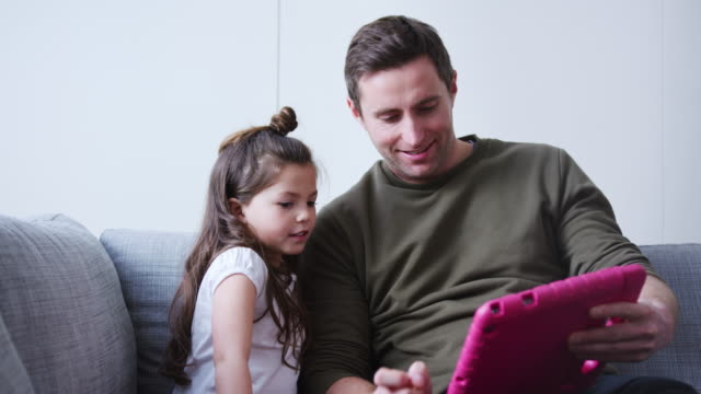 Padre-e-hija-sentados-en-el-sofá-en-casa-jugando-juntos-en-la-tableta-digital-en-el-caso-rosa-en-casa