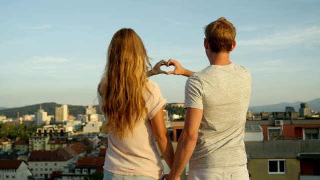 CLOSE-UP:-Man-and-woman-standing-on-rooftop-holding-hands-making-heart-symbol