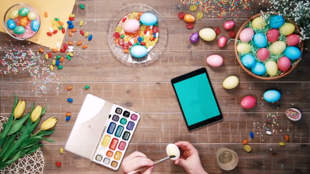 Man-painting-easter-egg-and-digital-tablet-with-green-screen-lies-on-table-decorated-with-easter-eggs-Top-view