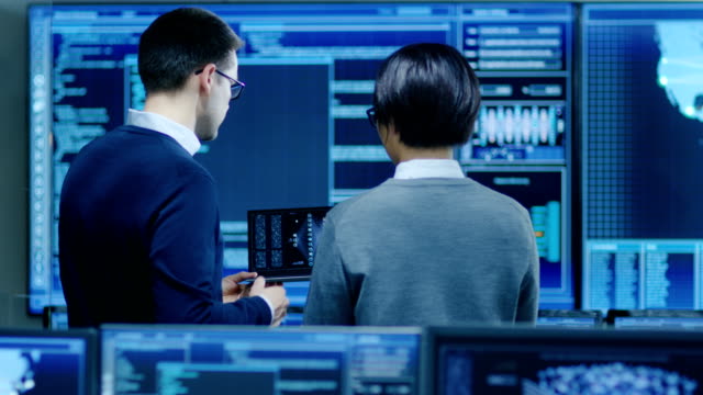 In-the-System-Control-Room-IT-Specialist-and-Project-Engineer-Have-Discussion,-they're-surrounded-by-Multiple-Monitors-with-Graphics.-They-Work-in-a-Data-Center-on-Data-Mining,-AI-and-Neural-Networking.