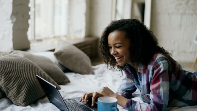 Lauging-rizado-afro-americano-niña-usando-laptop-para-compartir-redes-sociales-acostado-en-la-cama-en-casa
