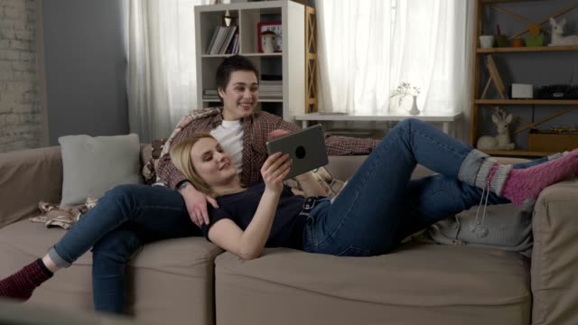 Lesbian-couple-is-resting-on-the-couch,-laughing-and-watching-funny-movie-on-tablet-60-fps