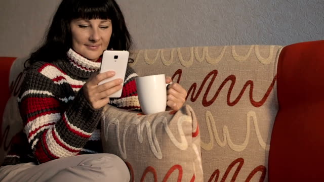 Young-woman-dressed-in-stripped-sweater-sits-on-sofa-,uses-smartphone,drinking-coffee.Girl-is-chatting,blogging,checking-email.-Online-marketing,-education,social-network.