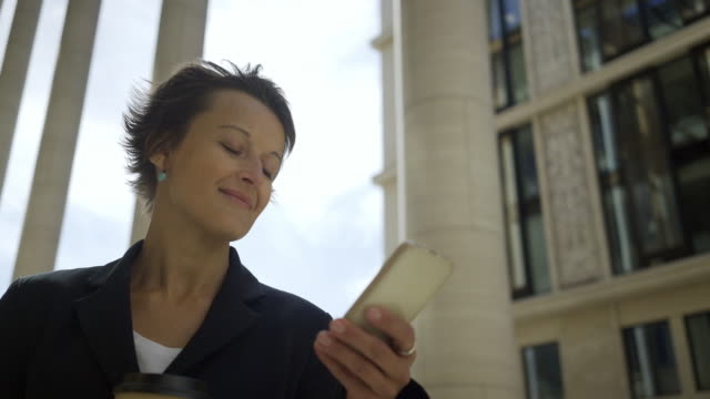 Schwenken-der-zuversichtlich-geschäftsfrau-mit-kurzen-Haaren-mit-Anwendung-auf-ihrem-Smartphone-im-Freien,-zum-Mitnehmen-Kaffeetasse-in-der-hand