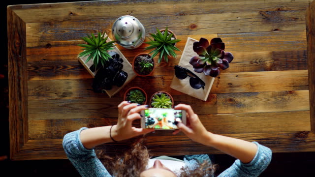 Creative-photographer-is-using-smartphone-to-take-flat-lay-pictures-of-plants,-camera-and-sunglasses-on-wooden-table,-woman-is-touching-screen-and-photographing.