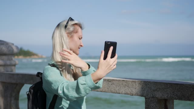 Junge-schöne-schlanke-Frau-mit-langen-blonden-Haaren-mit-Sonnenbrille-und-grünes-Hemd-stehen-in-der-Nähe-von-Palme-und-Selfie-auf-Handy-auf-blauem-Himmel-und-Meer-Hintergrund-zu-machen.-Mädchen-mit-smartphone