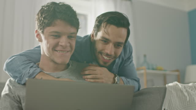 Entzückende-männliche-Gay-paar-verbringen-Zeit-zu-Hause.-Junger-Mann-arbeitet-auf-einem-Laptop,-seine-Partnerin-von-hinten-kommt-und-sanft-umarmt-ihn.-Sie-lachen-und-Hände-berühren.-Zimmer-ist-Modern-eingerichtet.