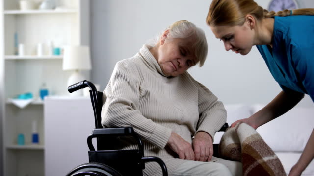 Young-nurse-covering-with-pled-old-woman-sleeping-in-wheelchair,-taking-care