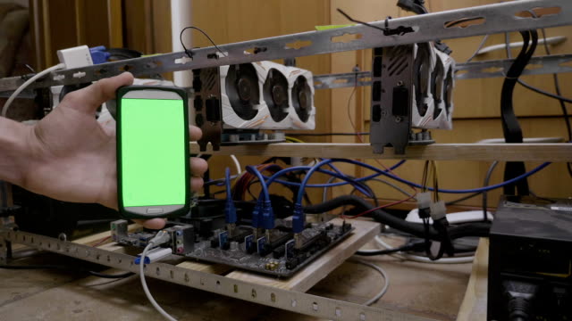 Programmer-engineer-showing-cellphone-with-green-screen-display-connecting-to-cryptocurrency-mining-rig-data