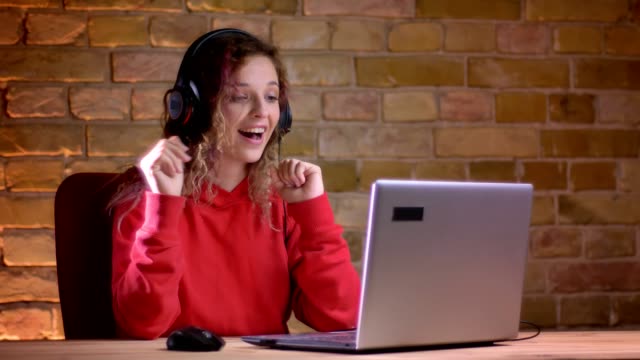 Portrait-of-young-female-blogger-in-red-hoodie-recording-video-using-laptop-on-bricken-wall-background.