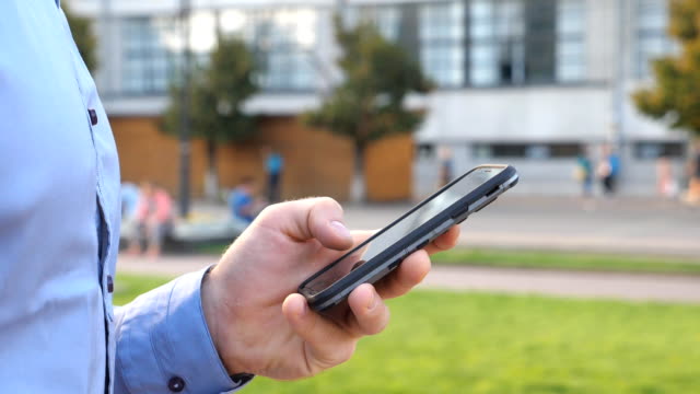 Close-up-hand-of-young-businessman-holding-and-touching-smartphone.-Unrecognizable-man-standing-on-urban-street-and-browsing-phone.-Guy-using-gadget-for-work.-Slow-motion-Side-view