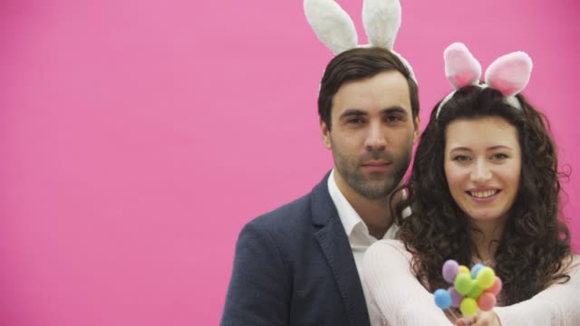 Young-creative-couple-on-pink-background.-With-hackneyed-ears-on-the-head.-During-this,-the-wife-holds-decorative-multicolored-eggs-in-her-hands.-Gently-kissing-her-husband-looking-at-the-camera-together.