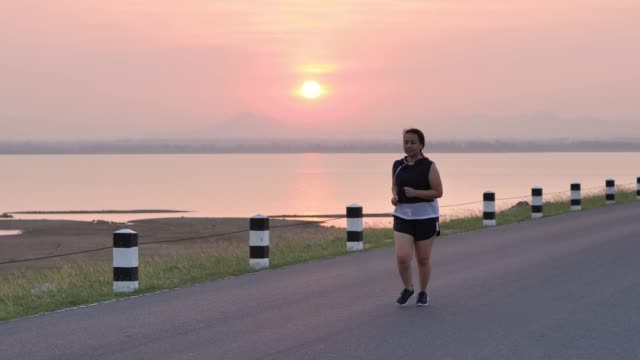 Übergewichtige-asiatische-Frauen-joggen-am-frühen-Morgen-im-Sonnenlicht-auf-der-Straße.-Konzept-des-Gewichtsverlustes-mit-Bewegung-für-die-Gesundheit.-die-Zeitlupe