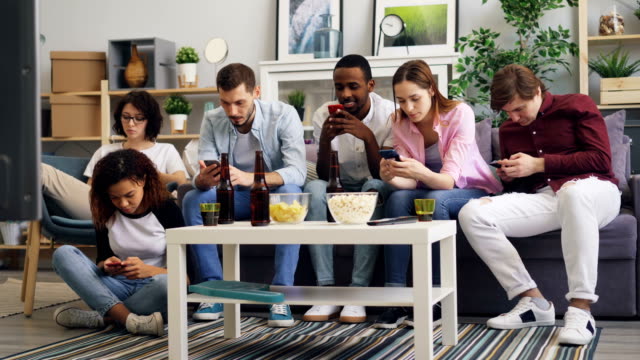 Multiracial-group-of-friends-using-smartphones-touching-screen-on-sofa-at-home