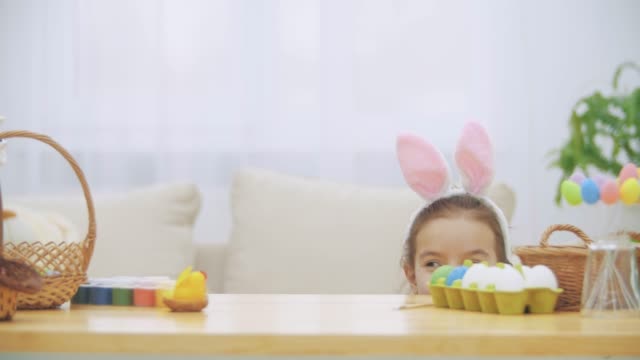 Nice-little-girl-with-bunny-ears-is-hiding-under-the-table-full-of-Easter-decorations.-Little-cute-white-bunny-is-attacking-girl's-head-kindly.-Playing-with-a-bunny-toy.