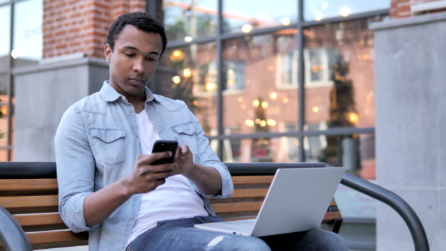 Afrikanischer-Mann-mit-Smartphone-und-Laptop,-Sitzen-auf-Bank