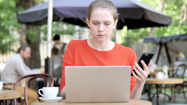 Young-Woman-Using-Smartphone-and-Laptop-Sitting-in-Cafe-Terrace
