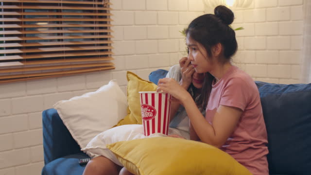 Lesbian-lgbt-women-couple-watching-movie-at-home,-Asian-female-lover-eating-popcorn-scary-and-shock-while-looking-horror-film-together-on-sofa-in-living-room-in-night-concept.