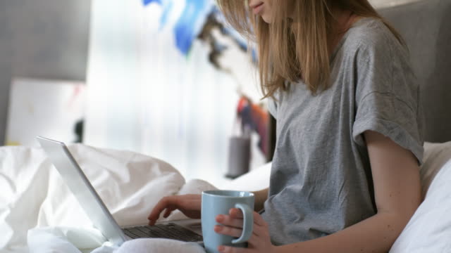 Frau-hält-Kaffee-und-Tippen-auf-Laptop-auf-dem-Bett