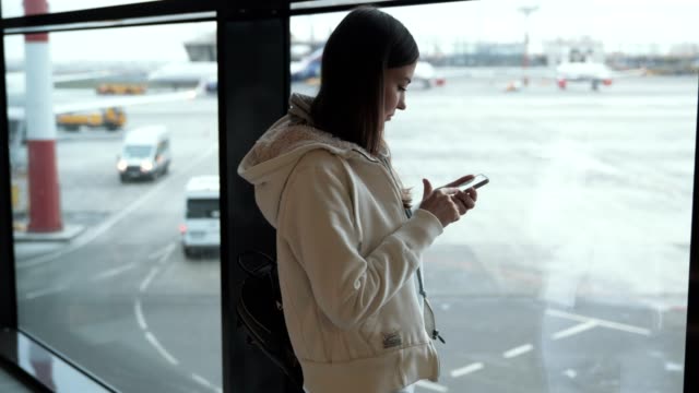 Woman-uses-mobile-phone-and-waves-her-hand-to-someone-from-window-at-airport