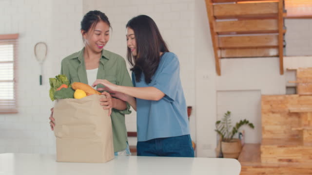 Asiatische-lesbische-Paar-halten-Lebensmittel-Einkaufspapiertüten-aus-dem-Supermarkt-setzen-sie-in-der-Küche-zu-Hause.