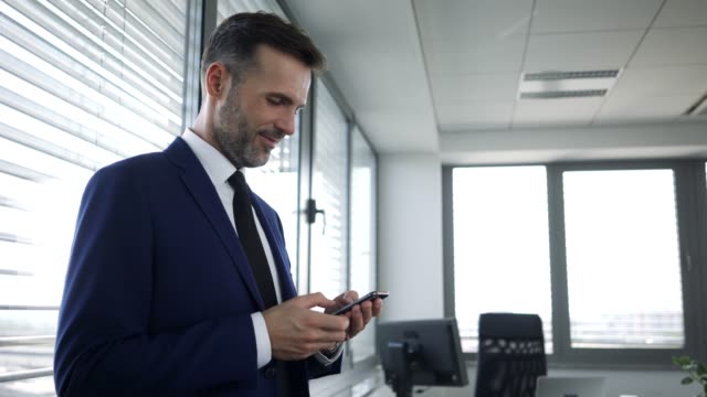 Happy-businessman-leaning-against-office-window-and-typing-on-smartphone
