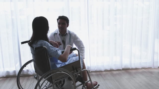 El-médico-es-el-aliento-y-la-empatía-asiática-de-las-mujeres-pacientes,-la-pierna-discapacitada-sentada-en-una-silla-de-ruedas-en-un-hogar-u-hospital-que-tiene-la-luz-del-sol-de-la-ventana.