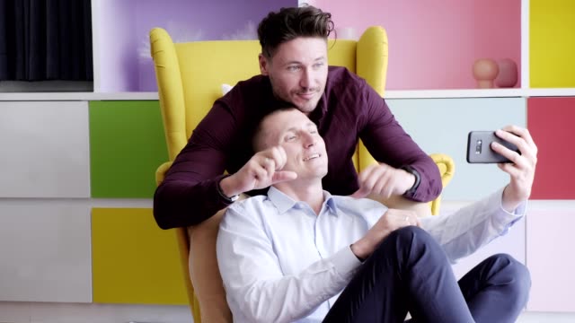 Schwules-Paar-mit-Telefon-auf-Sessel-zusammen.-Selfie-mit-Siegeszeichen.