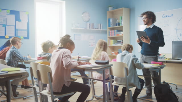 Begeisterter-Lehrer-verwendet-digitalen-Tablet-Computer,-während-er-einem-Klassenzimmer-voller-vielseitiger-heller-Kinder-eine-Lektion-erklärt.-In-der-Grundschule-Gruppe-von-Smart-Multiethnischen-Kinder-lernen