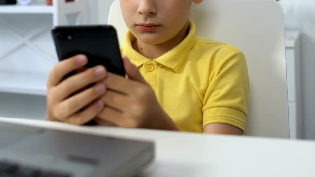 Niño-pequeño-sentado-frente-a-la-computadora-portátil,-viendo-vídeo-en-el-teléfono-inteligente,-adicción