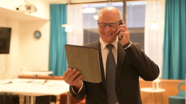 Feliz-hombre-de-negocios-senior-hablando-por-teléfono-mientras-usa-la-tableta-digital-en-el-trabajo
