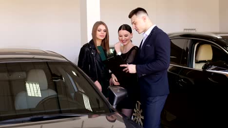 A-young-smiling-car-salesman-advises-two-girls-in-the-showroom.